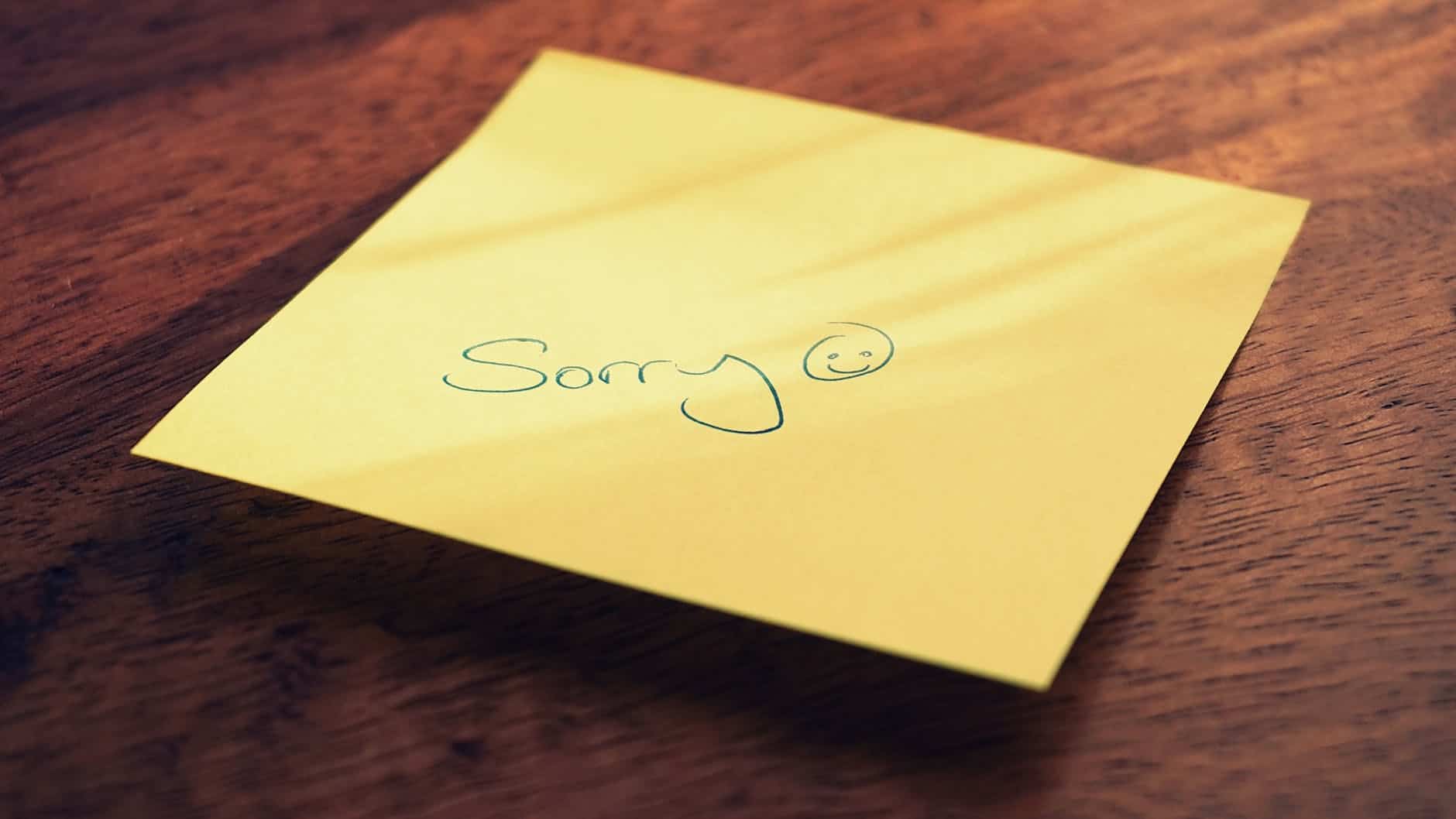 An apology note with a smiley face on a yellow sticky note on a wooden surface.
