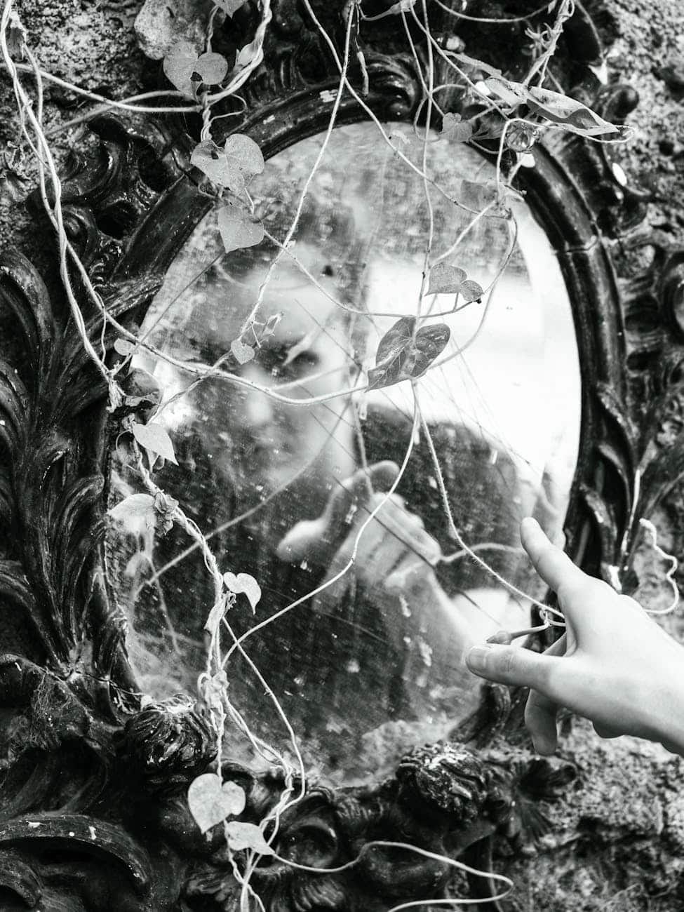 Person touching an old mirror with vines, symbolizing reflection and self-awareness.