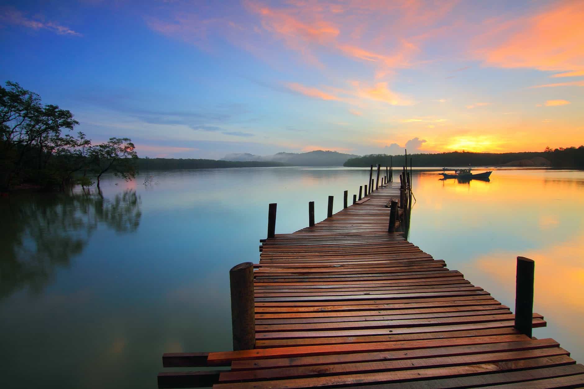 Sunset over calm lake with a wooden dock extending into the water, surrounded by trees and mountains.