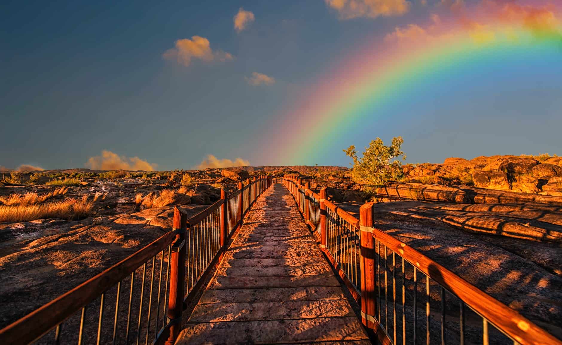 Rainbow arching over a wooden bridge in a desert with rocky terrain and a clear sky.