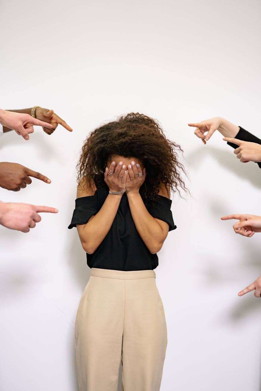 Woman covering her face surrounded by pointing fingers, representing emotional distress.